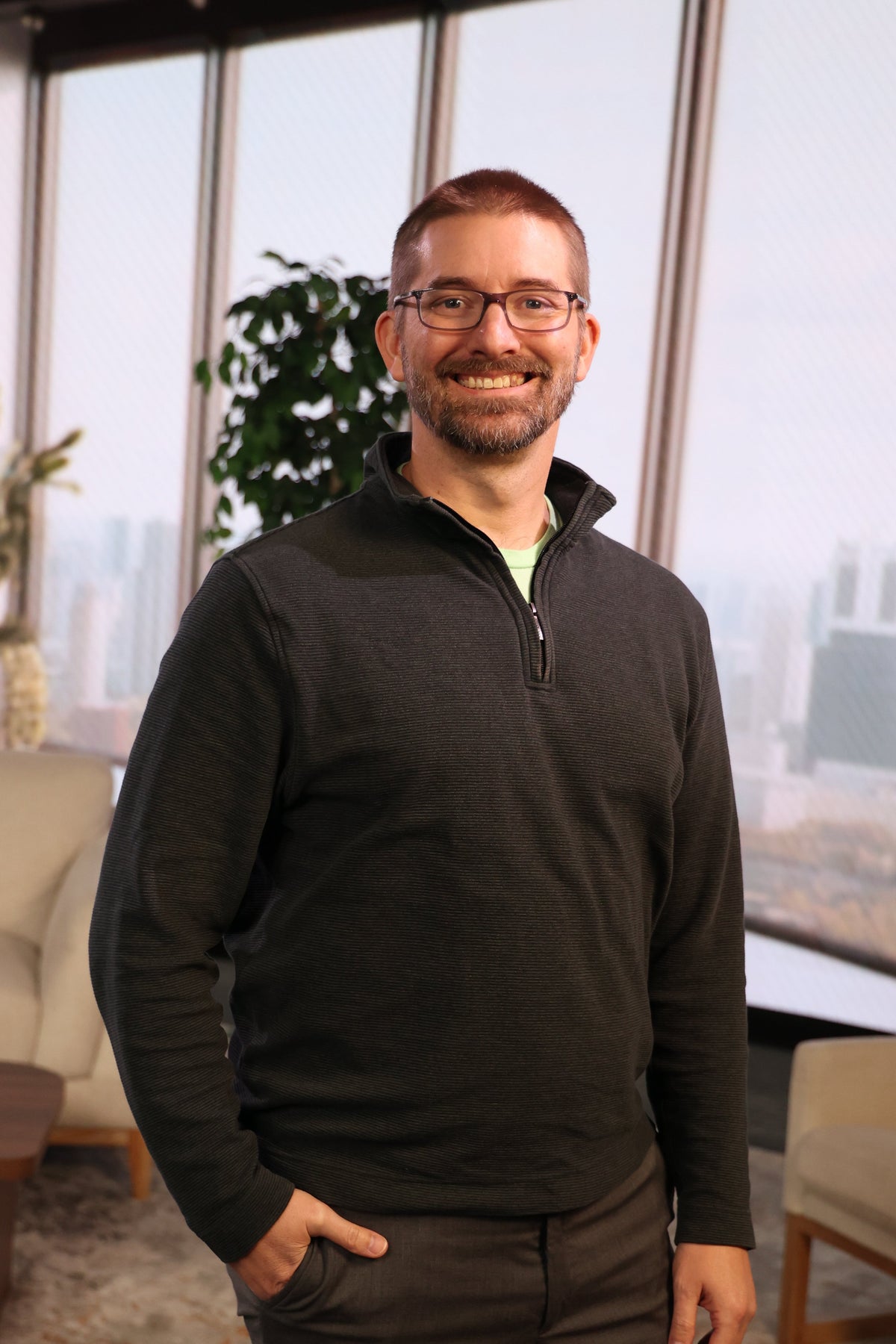 A smiling man with glasses and a beard stands indoors wearing a dark charcoal quarter-zip pullover sweater and dark pants, with a modern office or lounge setting featuring large windows, city views, and beige seating in the background.