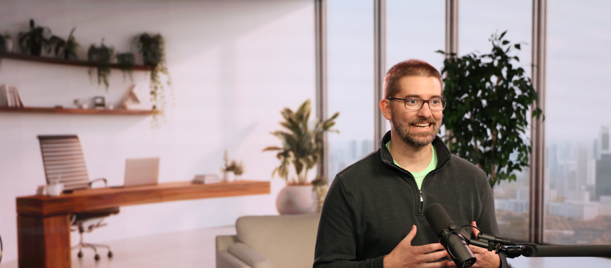 A man wearing glasses and a dark zip-up sweater stands in a modern office setting, holding a rifle with a scope. The room features a desk, office chair, sofa, and large windows overlooking a city skyline.